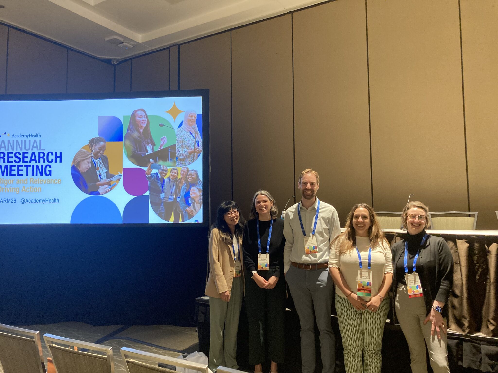 Tricia, Isabell, Ryan, Kat, and Vaughan standing on front of the Academy Health Annual Research Meeting slide and panelist table